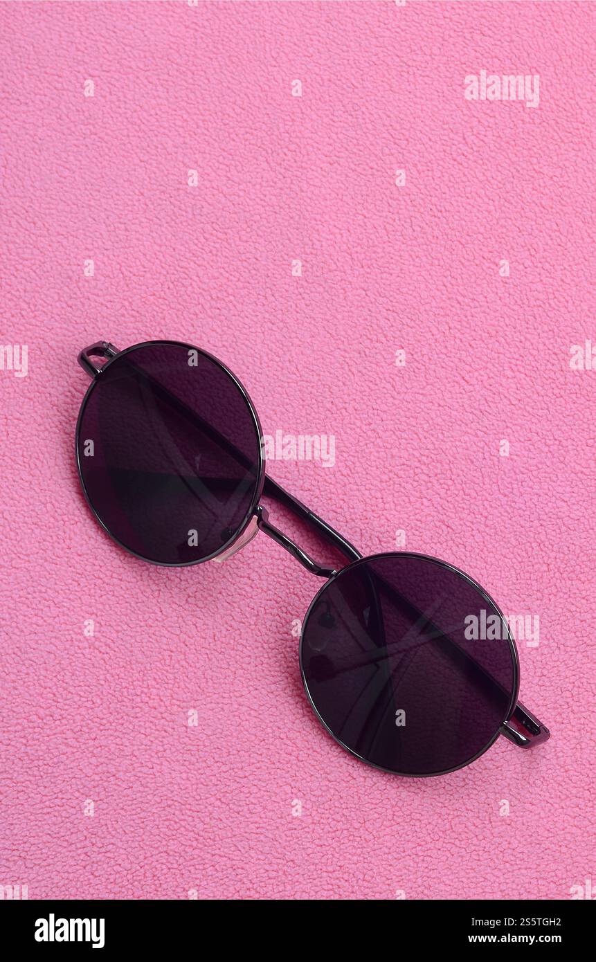 Gli eleganti occhiali da sole neri con occhiali rotondi si appoggiano su una coperta realizzata in morbido e soffice tessuto in pile rosa chiaro. Elegante immagine di sfondo in Foto Stock
