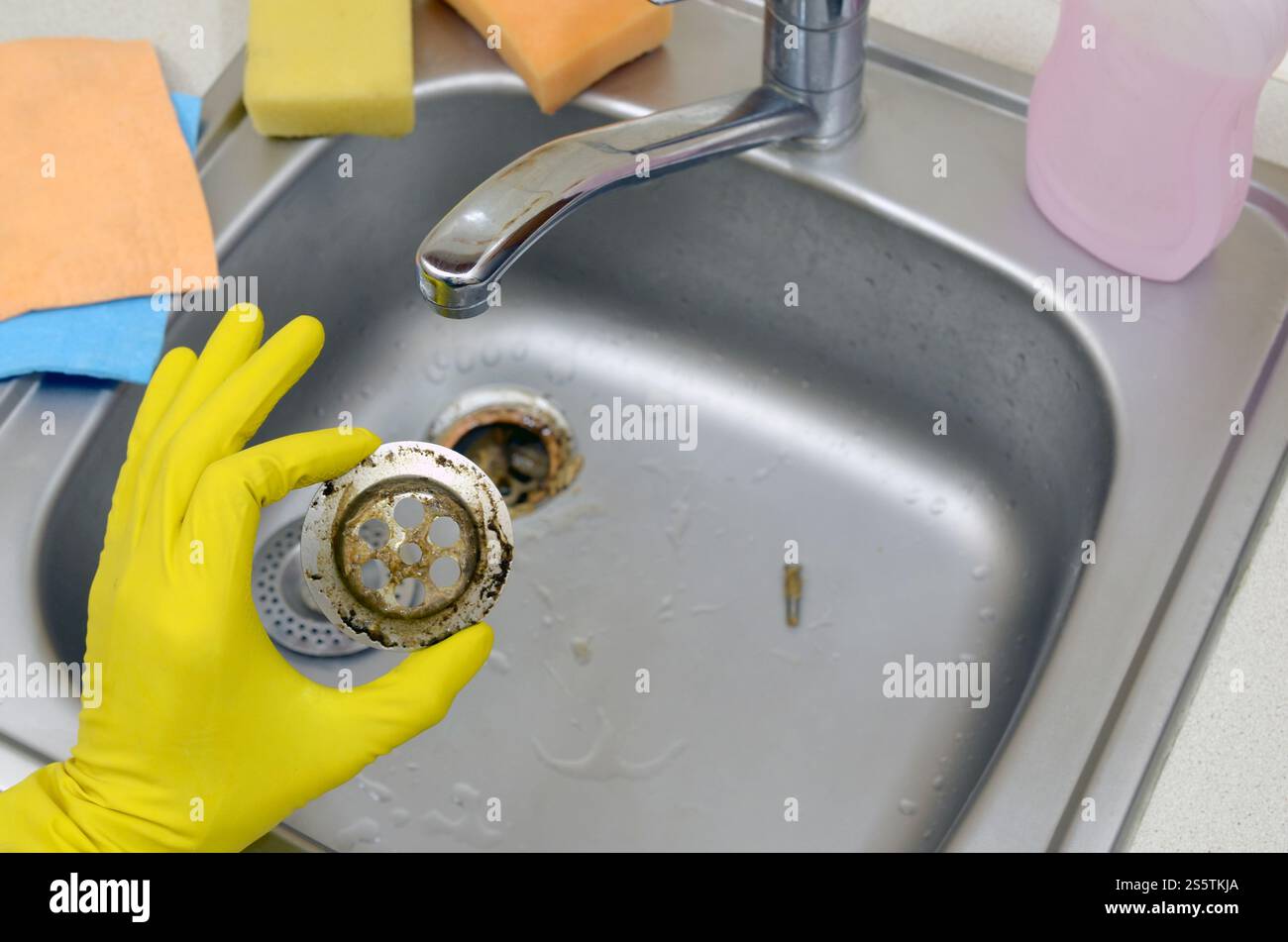 Il detergente nei guanti in gomma mostra i rifiuti nella protezione per peluche di un lavello da cucina. Filtro di scarico del lavandino in acciaio sporco. I rifiuti intrappolano la griglia dei fori di drenaggio. Foto Stock
