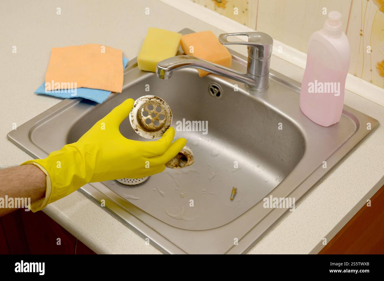 Il detergente nei guanti in gomma mostra i rifiuti nella protezione per peluche di un lavello da cucina. Filtro di scarico del lavandino in acciaio sporco. I rifiuti intrappolano la griglia dei fori di drenaggio. Foto Stock