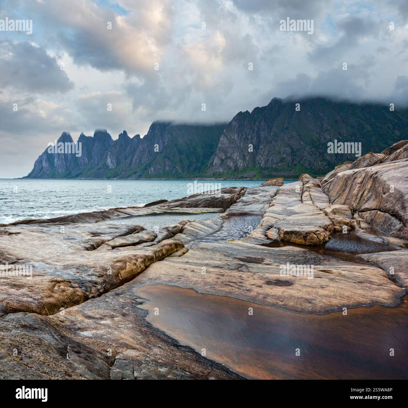 La spiaggia sassosa con bagni di marea a Ersfjord, Senja, Norvegia. Estate giorno polare notte costa. I denti di drago di roccia nel lontano. Foto Stock