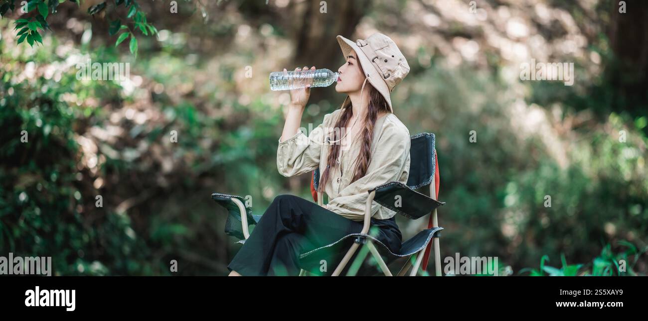 La giovane bella donna che indossa un cappello da campeggio e si siede su una sedia, beve acqua in bottiglia vicino al ruscello mentre campeggia nella foresta naturale, copia spazio Foto Stock