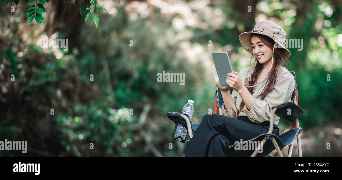 Giovane donna seduta su una sedia e utilizza una videochiamata con un tablet mentre campeggi nel parco naturale, spazio di copia Foto Stock