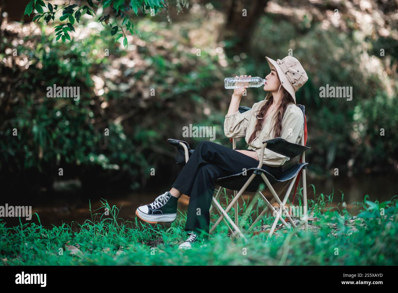 La giovane bella donna che indossa un cappello da campeggio e si siede su una sedia, beve acqua in bottiglia vicino al ruscello mentre campeggia nella foresta naturale, copia spazio Foto Stock