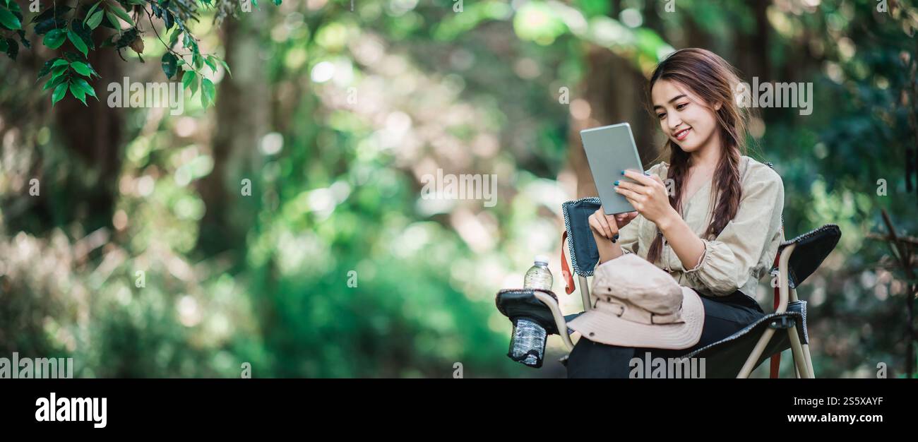 Giovane donna seduta su una sedia e utilizza una videochiamata con un tablet mentre campeggi nel parco naturale, spazio di copia Foto Stock