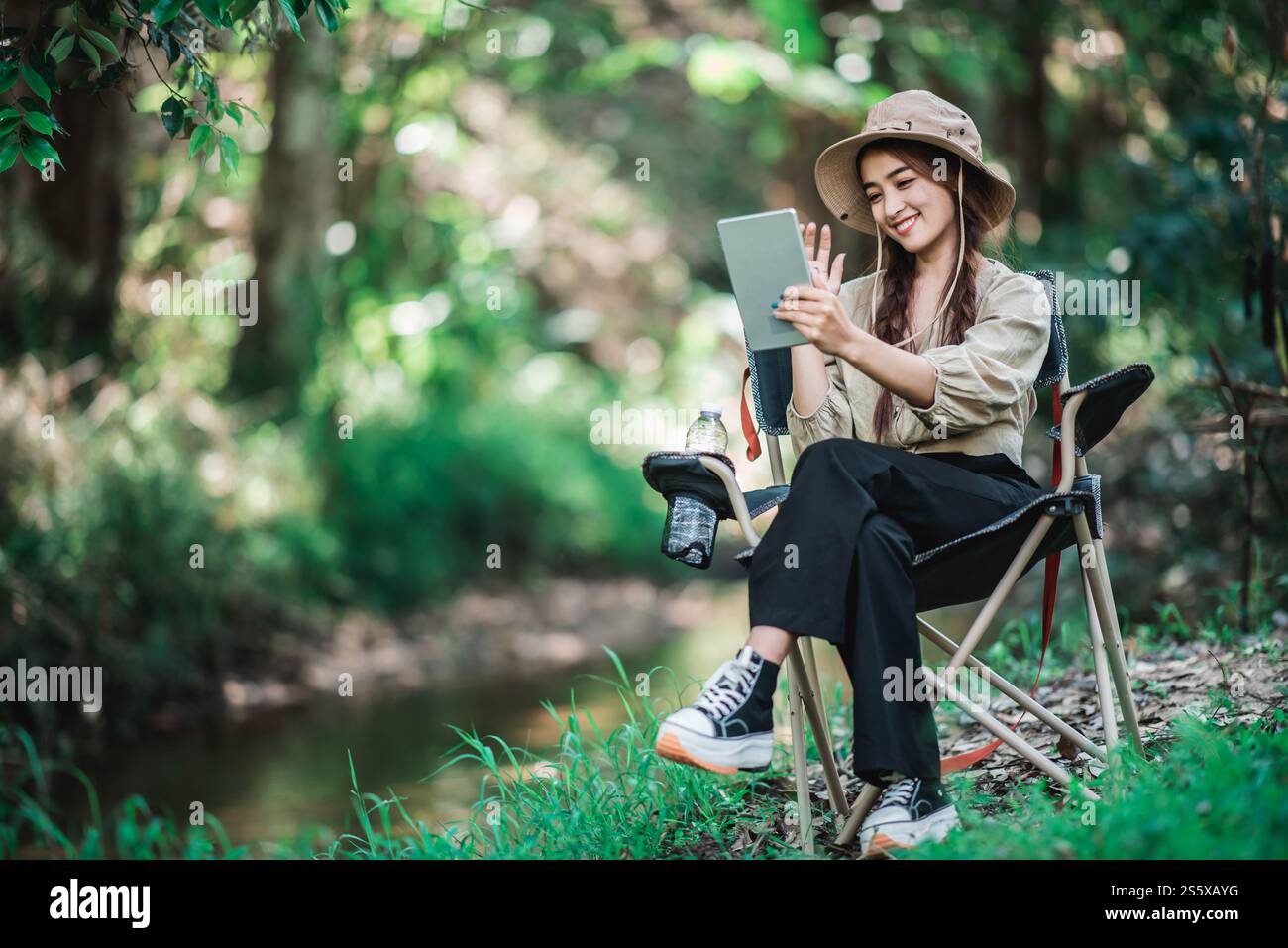 Giovane donna seduta su una sedia e utilizza una videochiamata con un tablet mentre campeggi nel parco naturale, spazio di copia Foto Stock