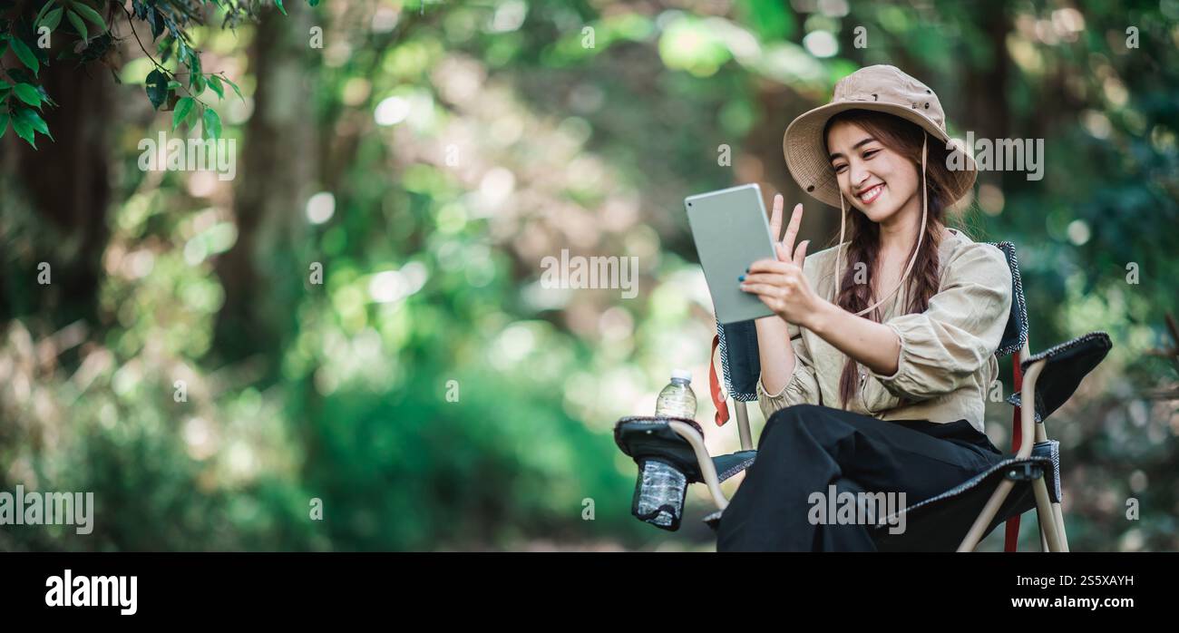 Giovane donna seduta su una sedia e utilizza una videochiamata con un tablet mentre campeggi nel parco naturale, spazio di copia Foto Stock