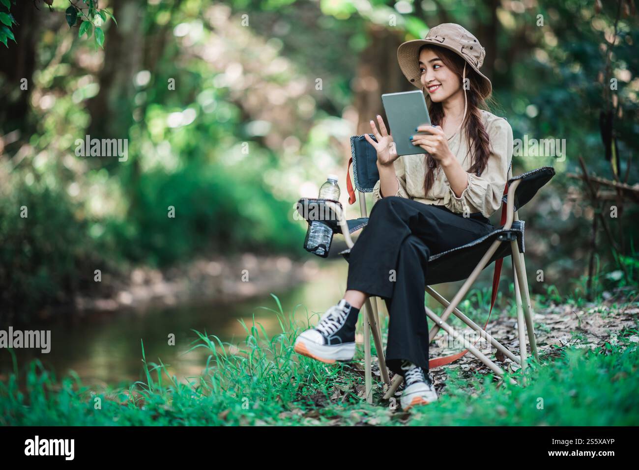 Giovane donna seduta su una sedia e utilizza una videochiamata con un tablet mentre campeggi nel parco naturale, spazio di copia Foto Stock