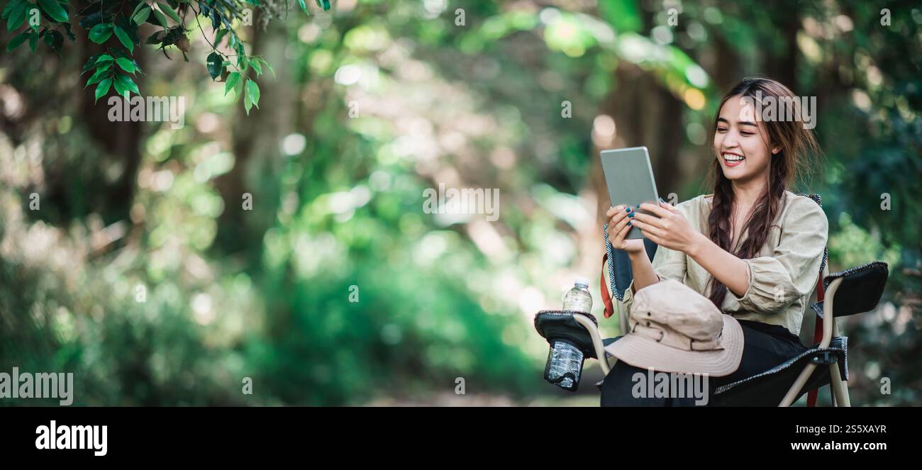 Giovane donna seduta su una sedia e utilizza una videochiamata con un tablet mentre campeggi nel parco naturale, spazio di copia Foto Stock