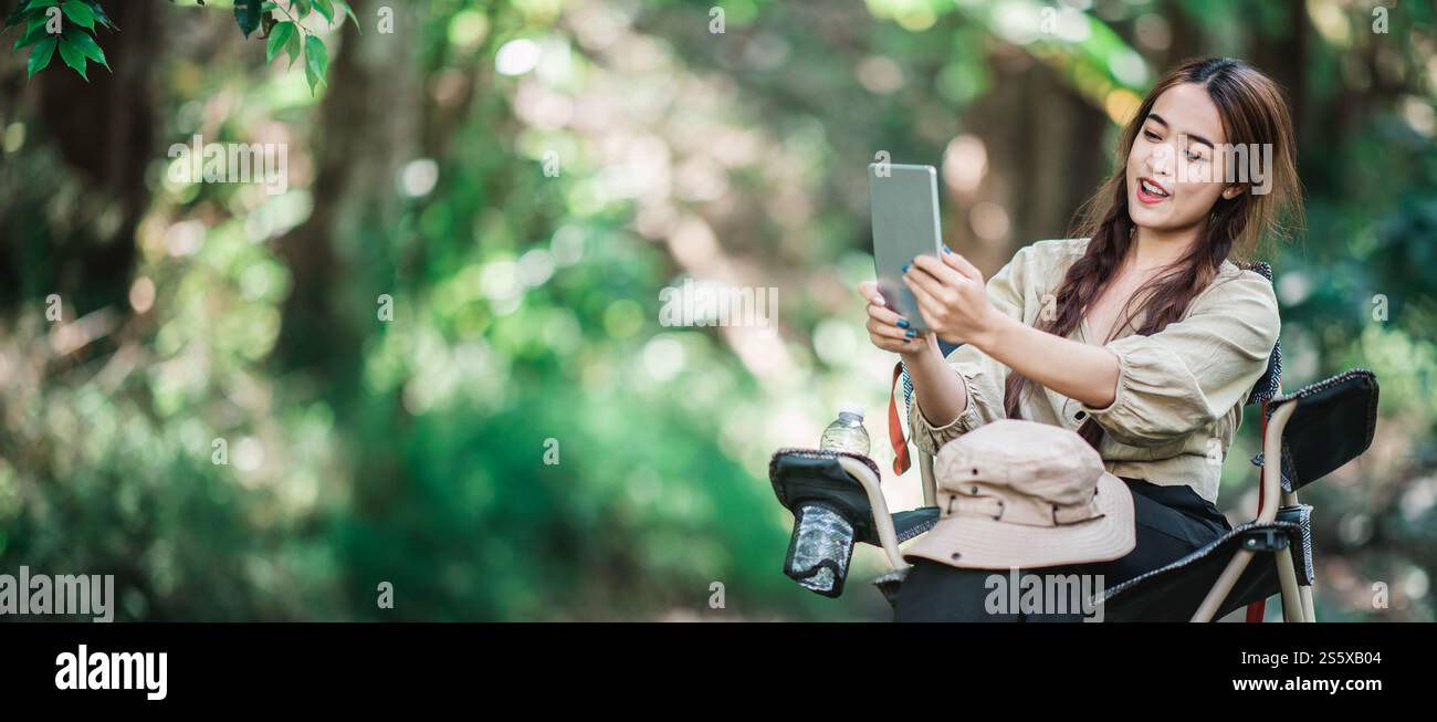 Giovane donna seduta su una sedia e utilizza una videochiamata con un tablet mentre campeggi nel parco naturale, spazio di copia Foto Stock