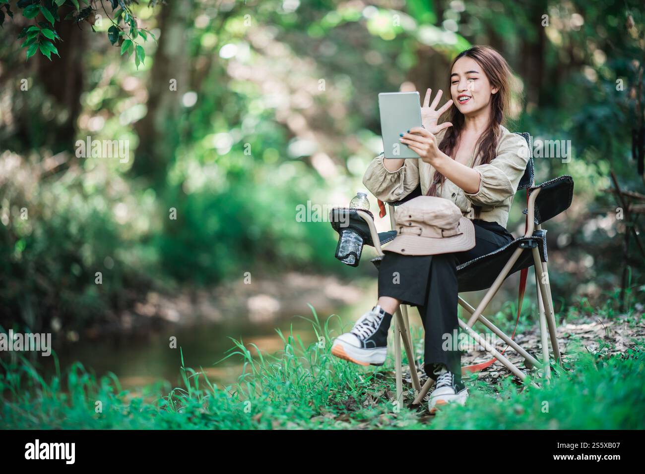 Giovane donna seduta su una sedia e utilizza una videochiamata con un tablet mentre campeggi nel parco naturale, spazio di copia Foto Stock