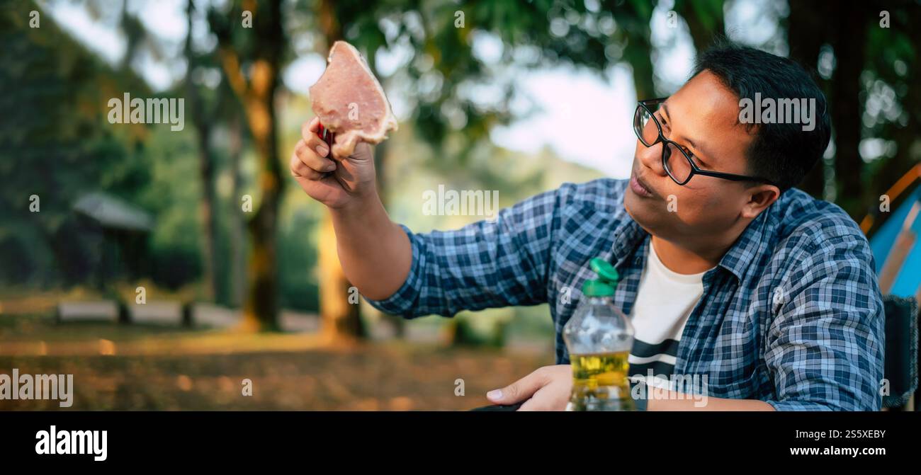 Ritratto dell'uomo viaggiatore asiatico che prepara una bistecca di maiale, barbecue in padella arrostita o pentola in campeggio. Cucina all'aperto, viaggi, campeggio, Foto Stock