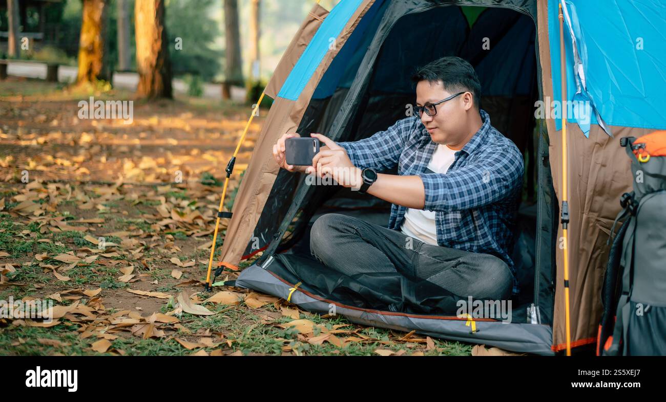 Ritratto dell'uomo viaggiatore asiatico occhiali che scattano una foto su smartphone in tenda campeggio. Viaggi all'aperto, campeggio e stile di vita. Foto Stock