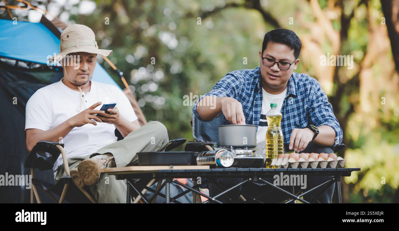 Ritratto di un uomo asiatico rilassato che usa lo smartphone e aspetta il suo amico che cucina con lo sfondo della tenda in campeggio. Piano cottura. All'aperto Foto Stock