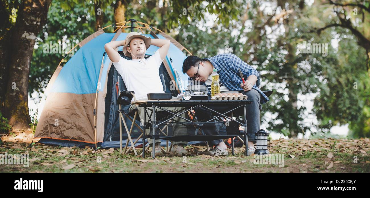 Uomo asiatico rilassato che usa lo smartphone e aspetta il suo amico che cucina con tenda in campeggio. Piano cottura. Cucina all'aperto, Foto Stock
