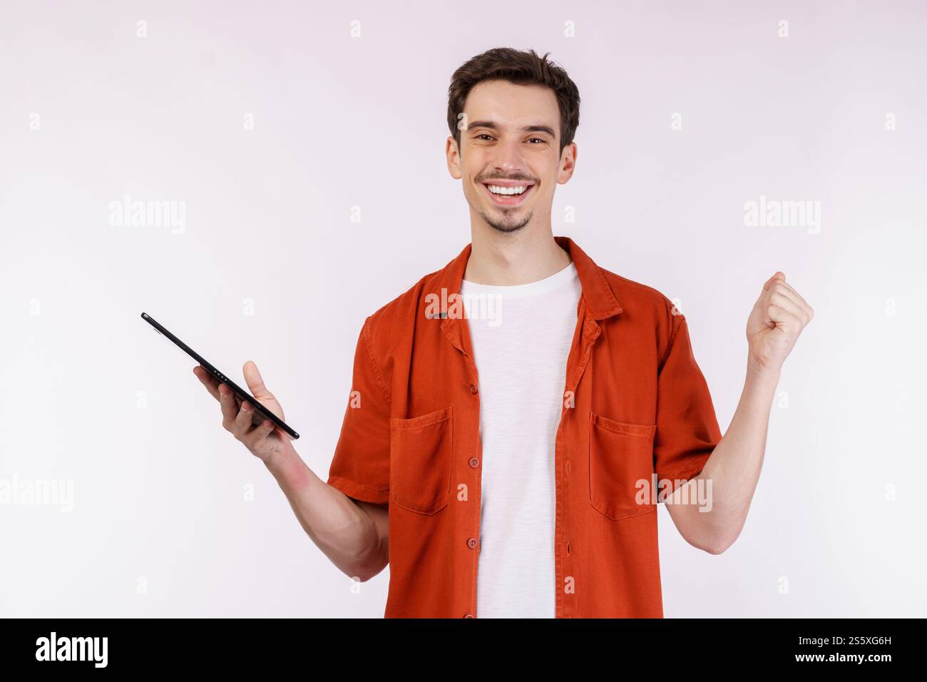 Ritratto di un uomo attraente e allegro che fa il gesto vincente stringendo il pugno e utilizzando l'app per dispositivi che cerca in rete isolata su sfondo bianco. Foto Stock