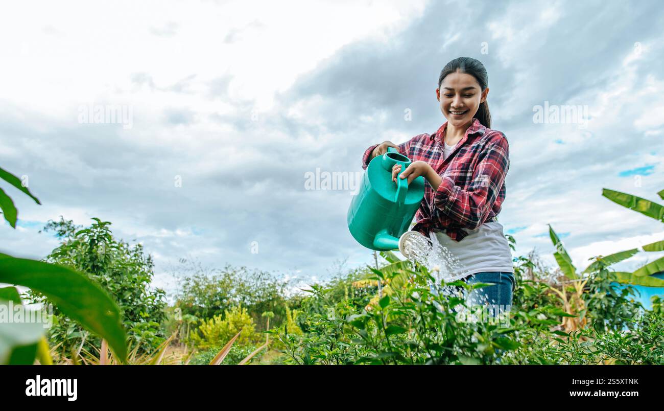 Giovane contadina asiatica che innaffia la pianta nel campo verde. Tecnologie moderne nella gestione dell'agricoltura e nel concetto di agroalimentare. Foto Stock