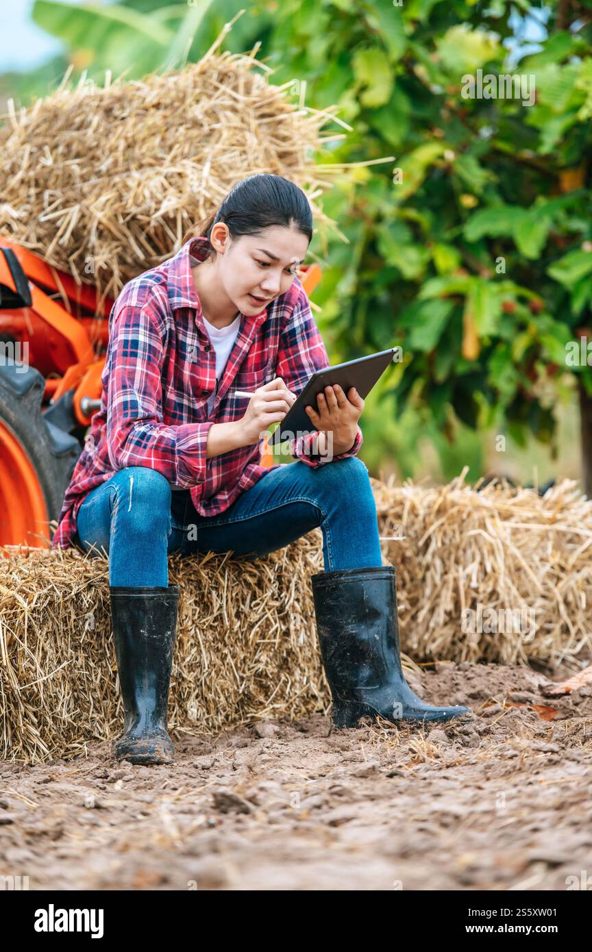 Giovane contadina asiatica seduto su una balla di fieno con una grande macchina trattore in campagna. Tecnologie moderne nella gestione dell'agricoltura e nel settore agroalimentare Foto Stock