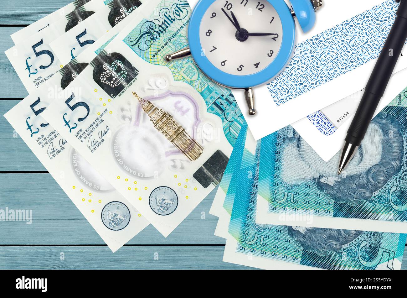 Banconote da 5 sterline inglesi e sveglia con penna e buste. Concetto di stagione fiscale, termine di pagamento per credito o prestito. Operazioni finanziarie utilizzando Foto Stock