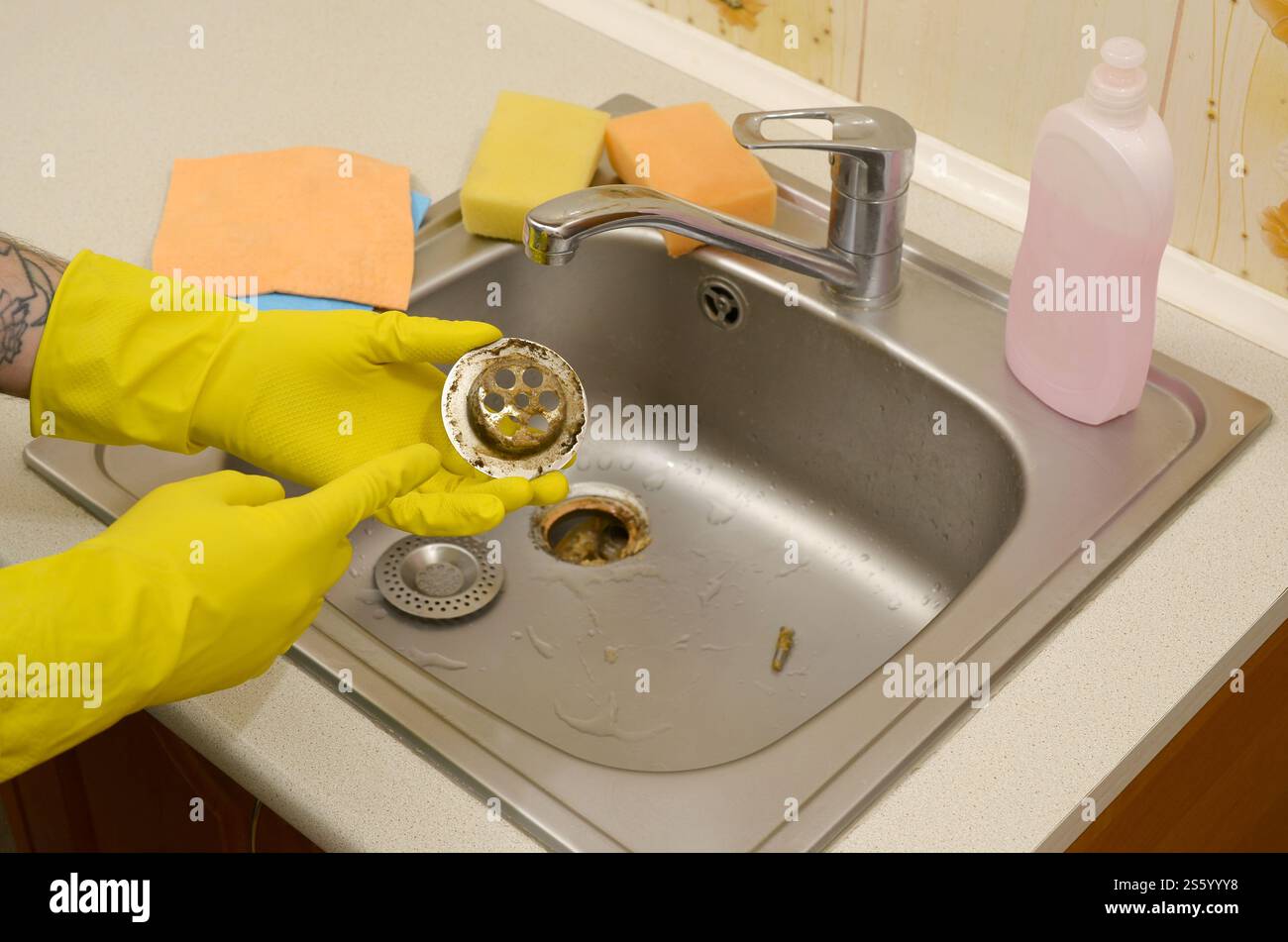 Il detergente nei guanti in gomma mostra i rifiuti nella protezione per peluche di un lavello da cucina. Filtro di scarico del lavandino in acciaio sporco. I rifiuti intrappolano la griglia dei fori di drenaggio. Foto Stock