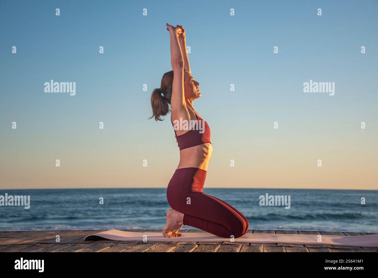 donna inginocchiata su un tappetino da yoga con le braccia sollevate, meditando all'alba o al tramonto sull'oceano Foto Stock