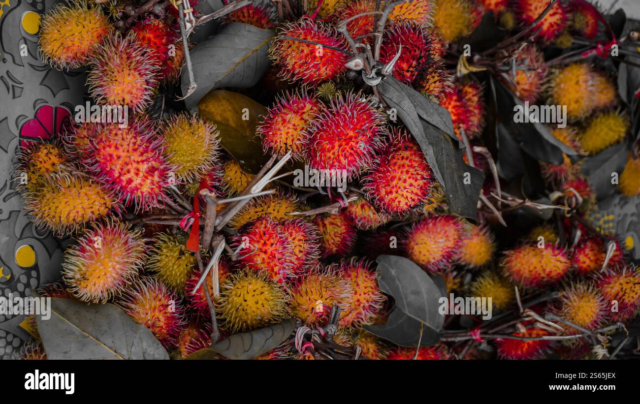 Una pila di frutti di rambutan freschi con vivaci pelli rosse e gialle, circondate da foglie verdi, esposte su una superficie a motivi geometrici. Foto Stock