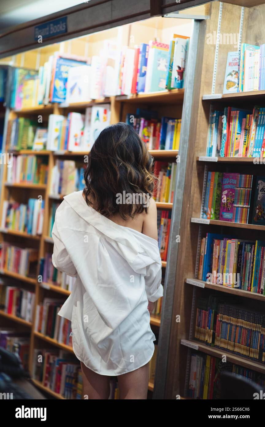 Una ragazza dai capelli scuri con una camicia bianca sta guardando un muro di libri in una libreria Foto Stock