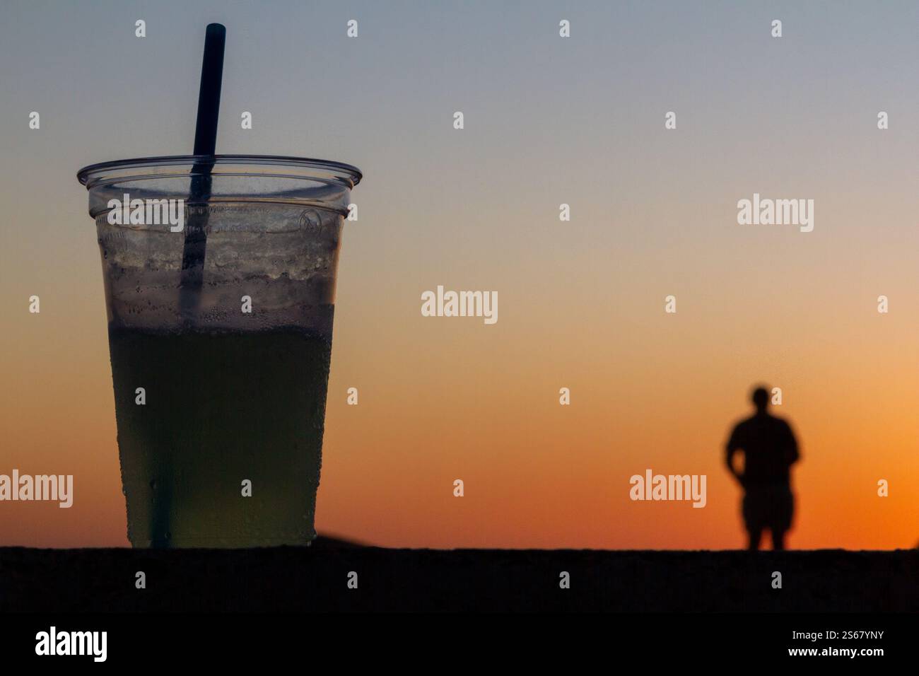 Cocktail sagomato in una tazza di plastica con ghiaccio e paglia contro un vivace tramonto arancione, con una figura sfocata che passeggia sulla spiaggia. Foto Stock Cocktail sagomato in una tazza di plastica con ghiaccio e paglia contro un vivace tramonto arancione, con una figura sfocata che passeggia sulla spiaggia. Foto Stock