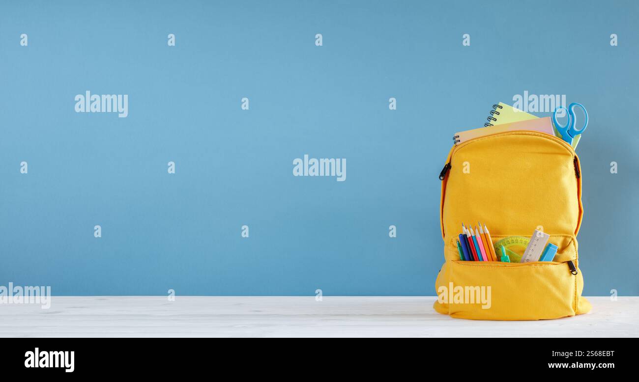 Uno zaino giallo con un muro blu dietro di esso. lo zaino è pieno di materiale scolastico come matite, penne e forbici. Torna all'organo scolastico Foto Stock