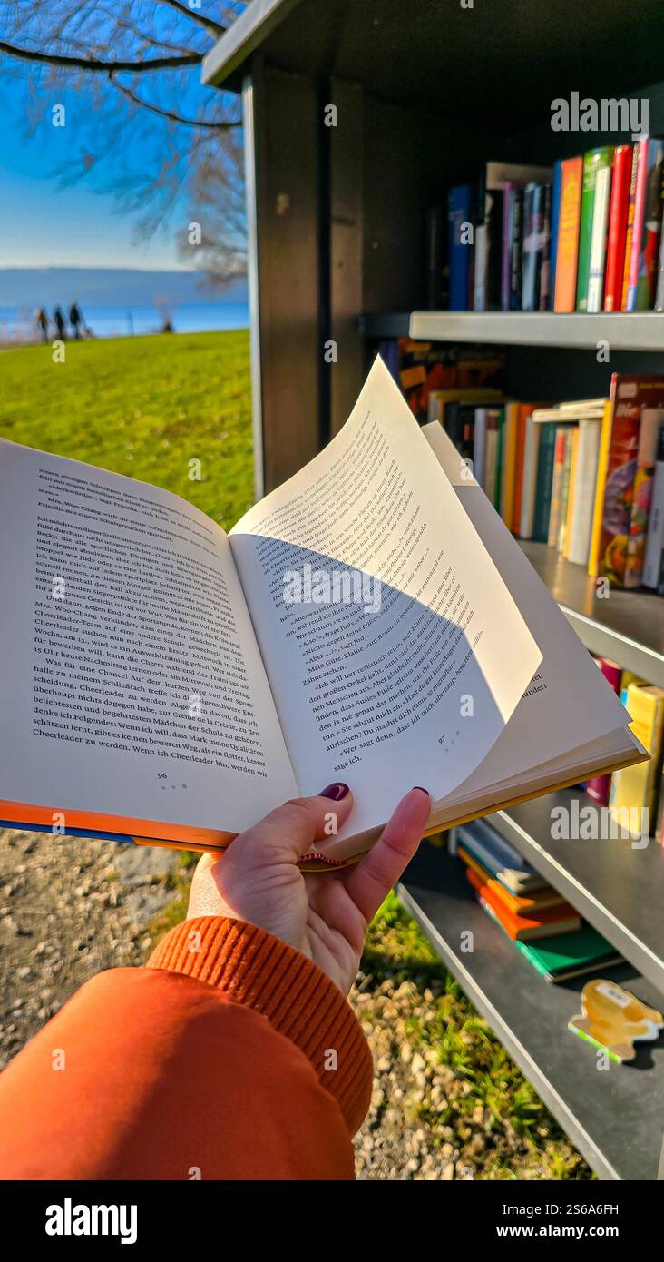 La mano delle donne tiene il libro aperto sullo sfondo di una biblioteca pubblica aperta gratuita per il bookcrossing nel parco cittadino. 26 dicembre 2024 Foto Stock