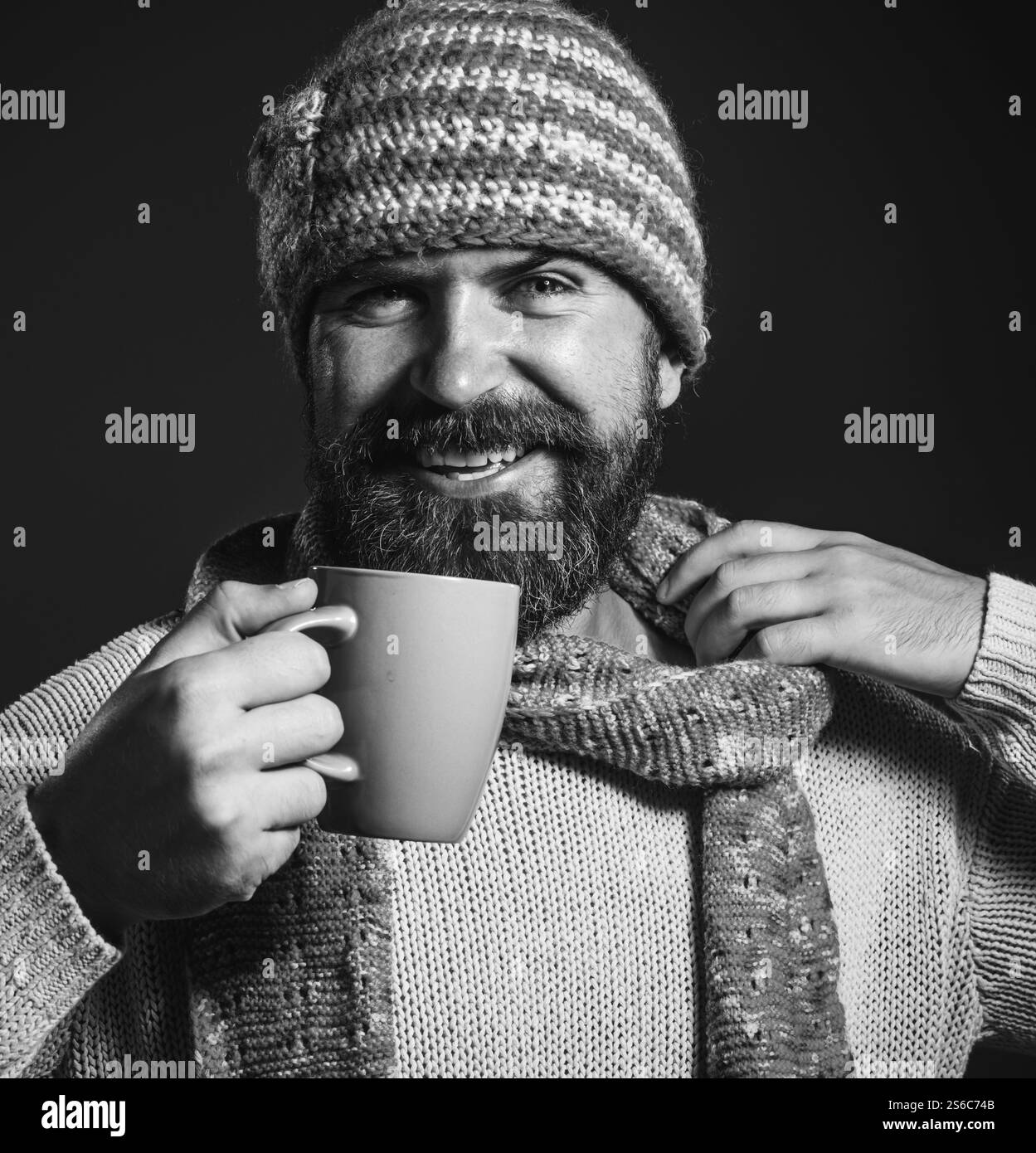 L'ora del caffè. Ritratto ravvicinato di un uomo barbuto sorridente con cappello a maglia, sciarpa e maglione bevendo una tazza di tè o caffè. Bell'uomo con barba in casua Foto Stock