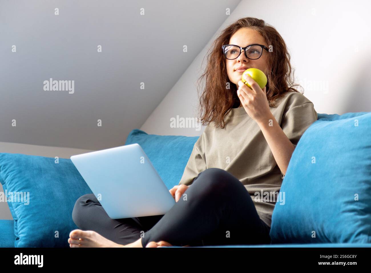 Donna che mangia mela verde mentre usa il suo laptop seduto sul divano di casa. Foto Stock