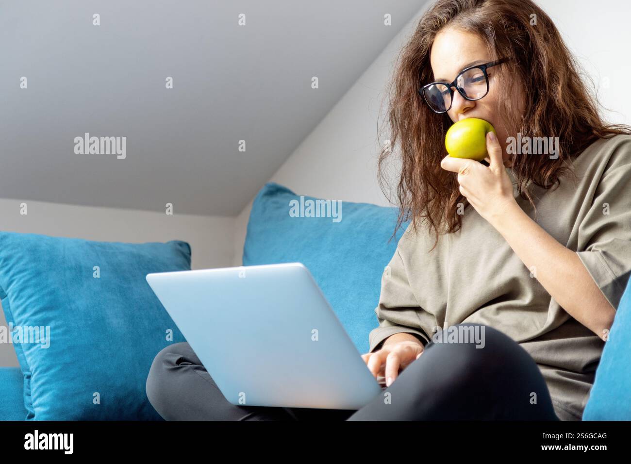 Donna che mangia mela verde mentre usa il suo laptop seduto sul divano di casa. Foto Stock