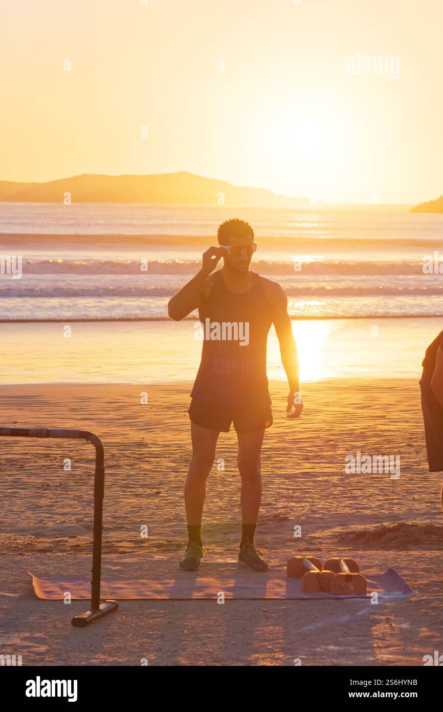 Il giovane atletico si trova accanto alle attrezzature sportive sulla spiaggia mentre le onde si infrangono al tramonto con l'isola alle spalle. Essaouira, Marocco. Gennaio 2025 Foto Stock