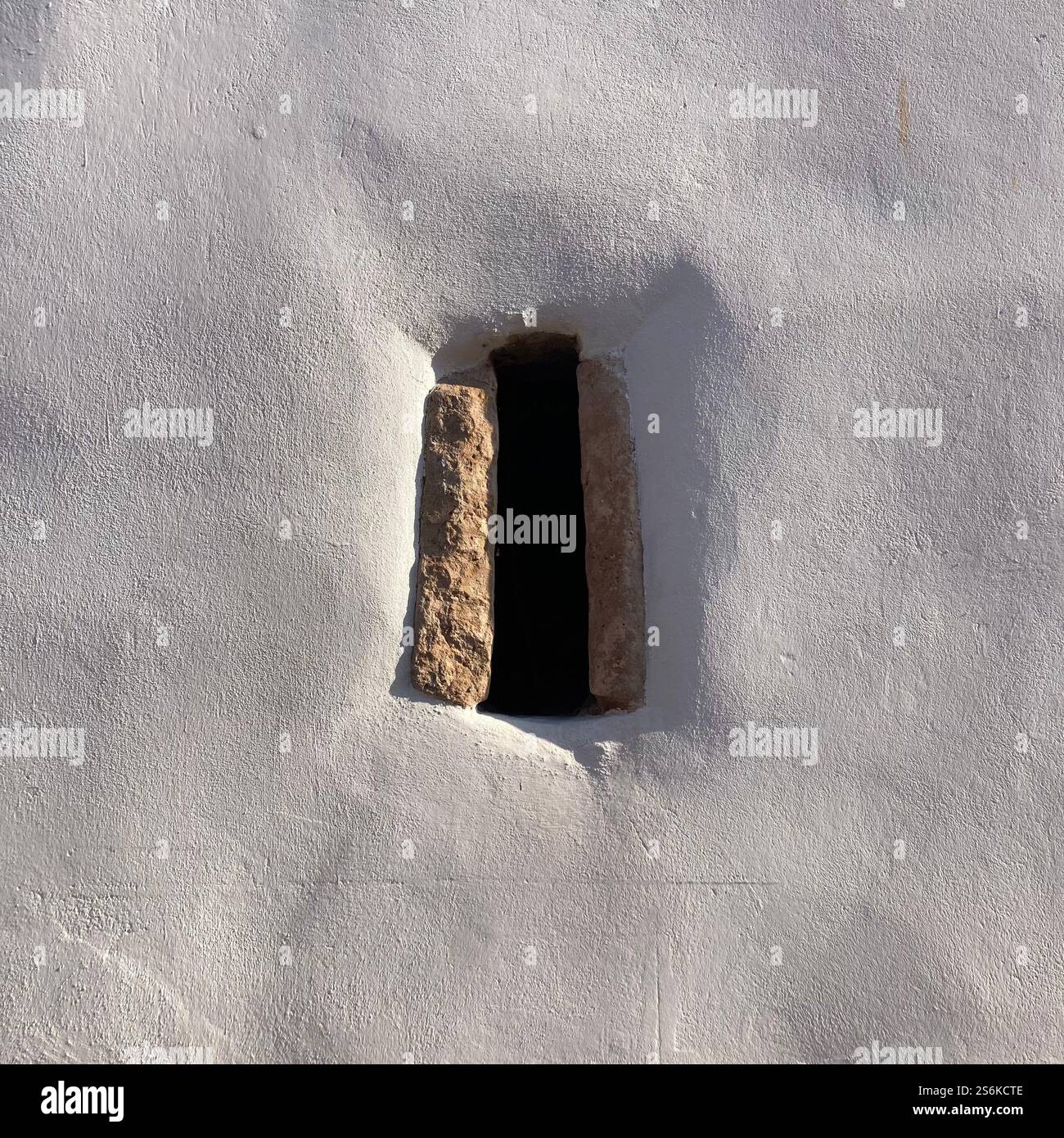Dettaglio di una finestra stretta con una cornice in pietra contro una parete in gesso bianco testurizzato, che mostra un design rustico senza tempo e un contrasto di illuminazione naturale Foto Stock