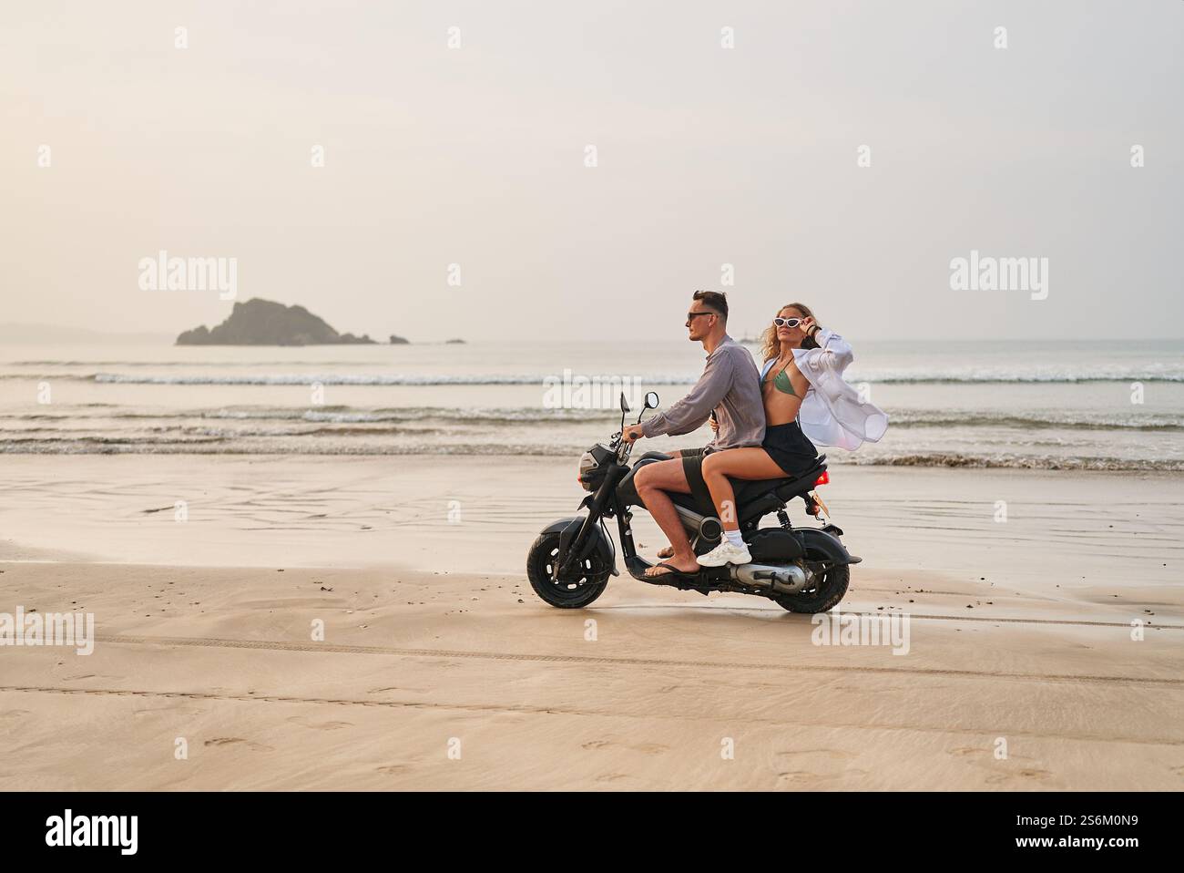 Due giri in moto lungo la costa sabbiosa. L'uomo con occhiali da sole guida, la donna in bikini ama la brezza. Viaggi avventurosi. Vista sulla spiaggia, onde sull'oceano Foto Stock