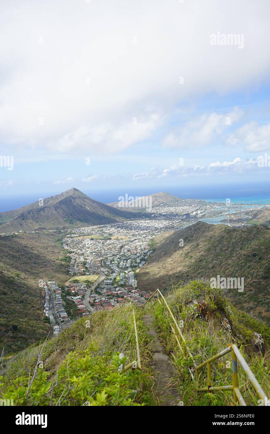 Vista panoramica da un sentiero escursionistico su Oahu, Hawaii, che si affaccia su vallate lussureggianti, aree urbane e il turchese Oceano Pacifico sotto un cielo parzialmente nuvoloso Foto Stock