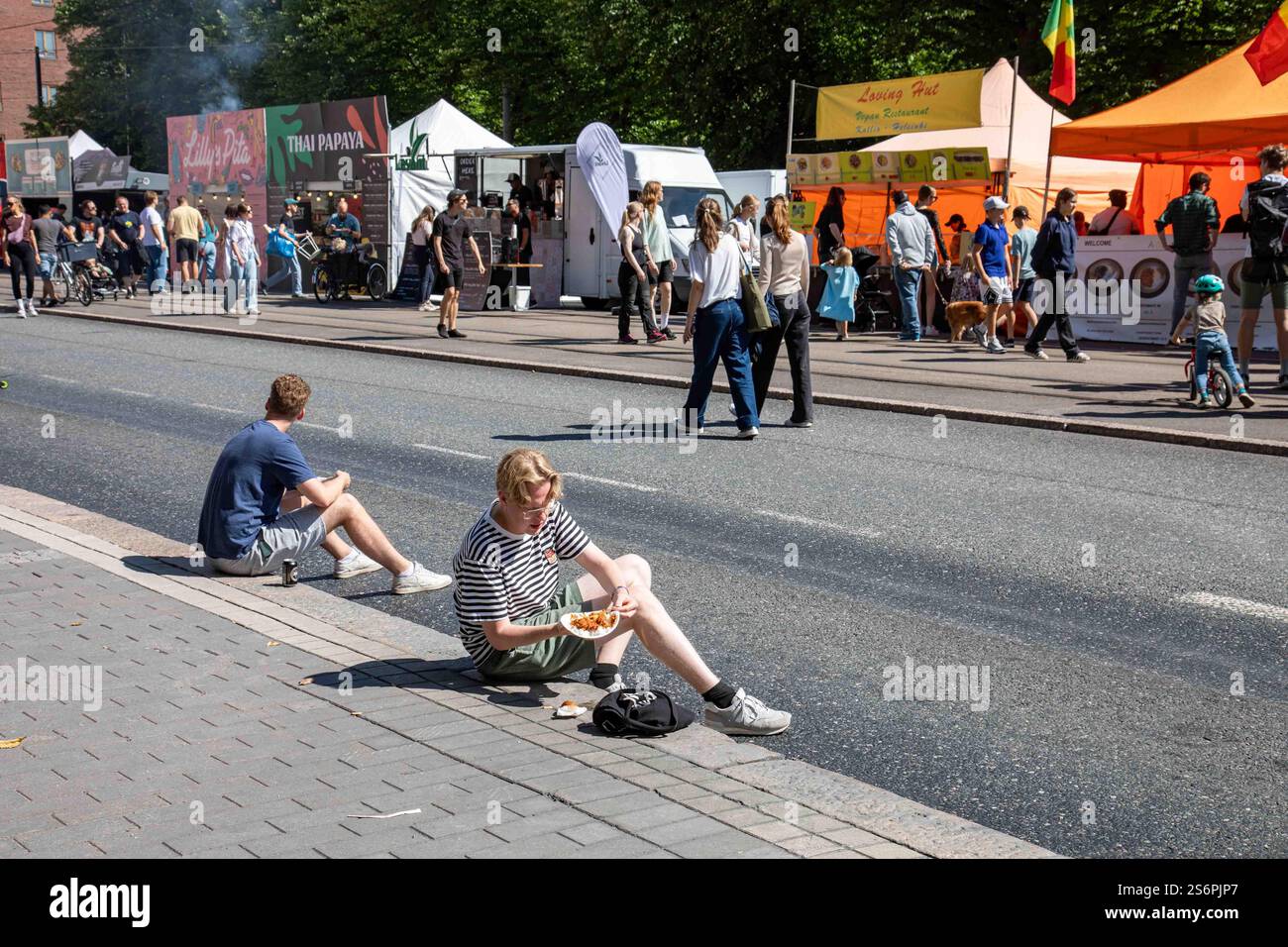 Le persone sedute sul marciapiede mangiano cibo di strada con una linea di venditori di cibo di strada sullo sfondo al Kallio Block Party 2024 di Helsinki, Finlandia Foto Stock