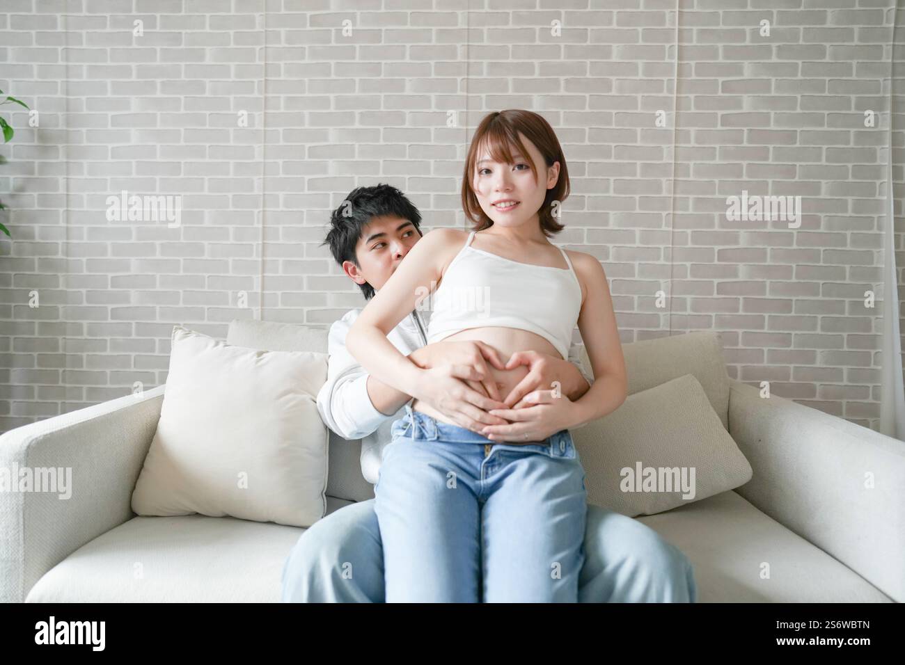 Una donna incinta giapponese di vent'anni, vestita per mostrare il suo stomaco, si siede su un divano nel suo appartamento, e suo marito la tiene, e fanno un cuore W Foto Stock