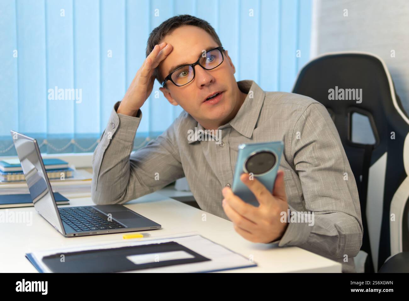uomo d'affari premuroso con una camicia blu che tiene in mano un telefono e guarda con attenzione lo schermo. Un giovane sta leggendo attentamente messaggi su una smartpho Foto Stock