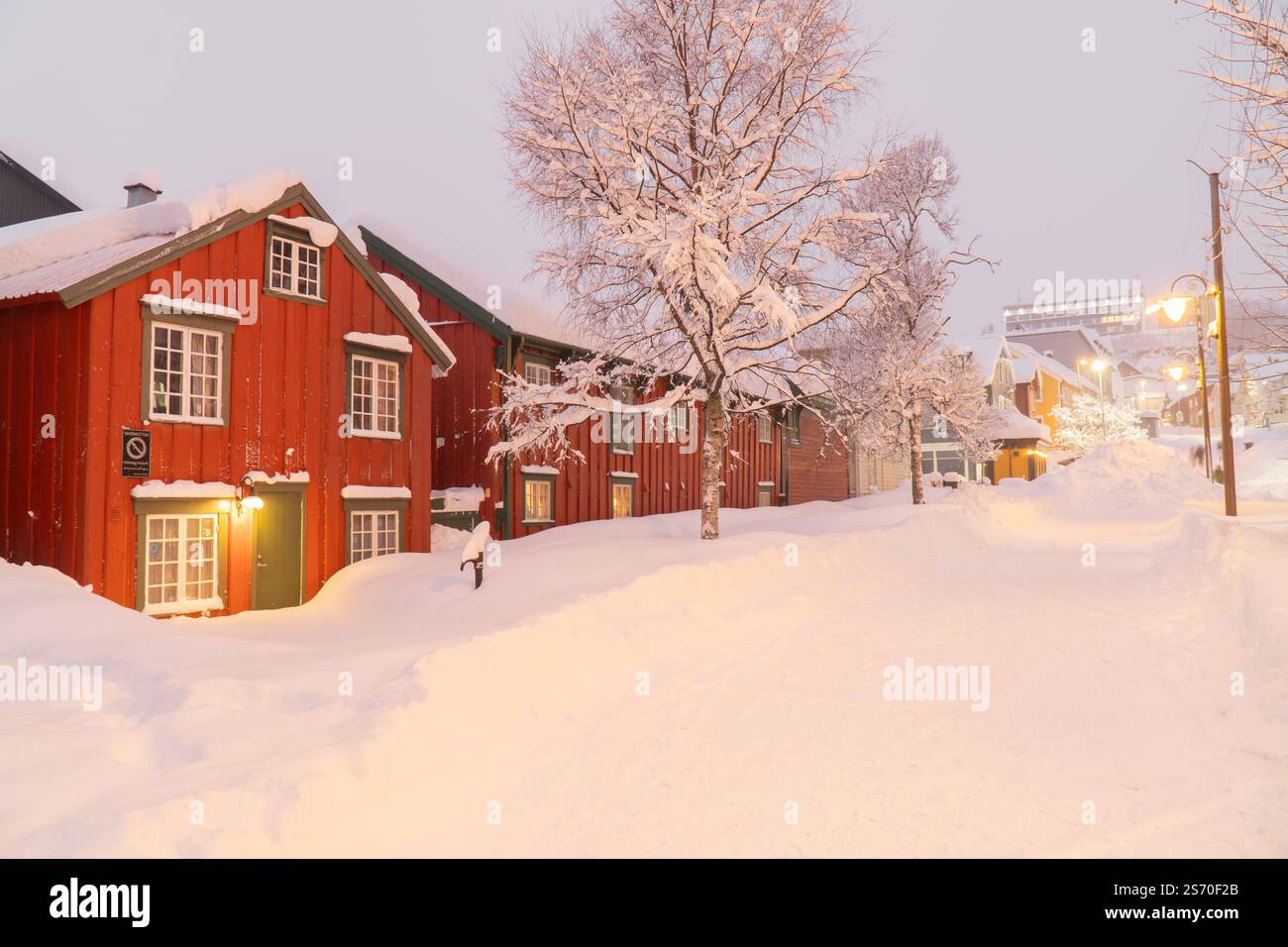 Case storiche in legno rosso e alberi innevati costeggiano una strada tranquilla sepolta nella neve invernale profonda durante un crepuscolo soffuso a Tromsø, Norvegia Foto Stock