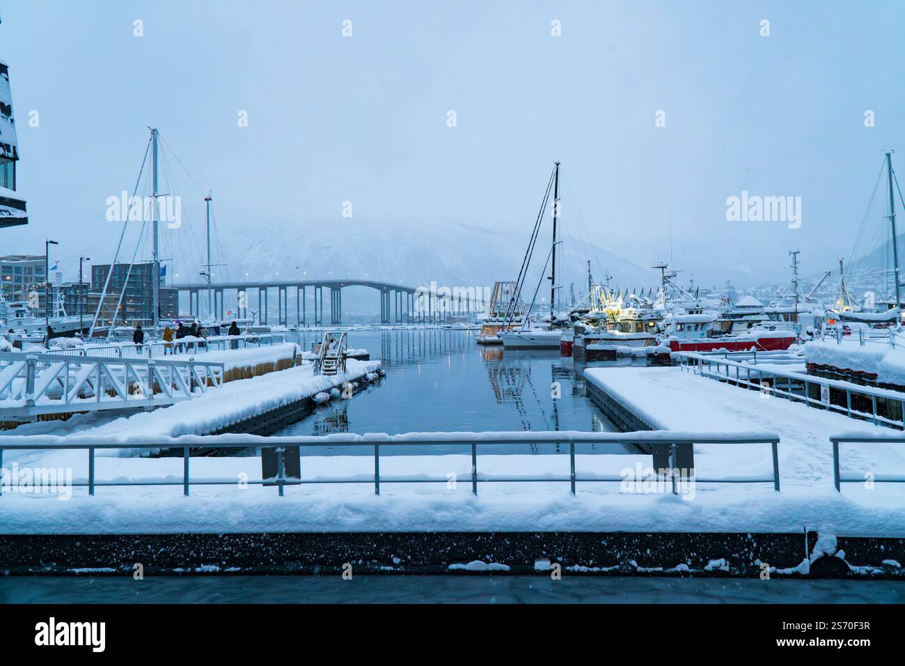 Moli del porto innevati con barche e yacht ormeggiati che si affacciano sul ponte distante di Tromso sotto un cielo invernale nuvoloso a Tromso, Norvegia Foto Stock