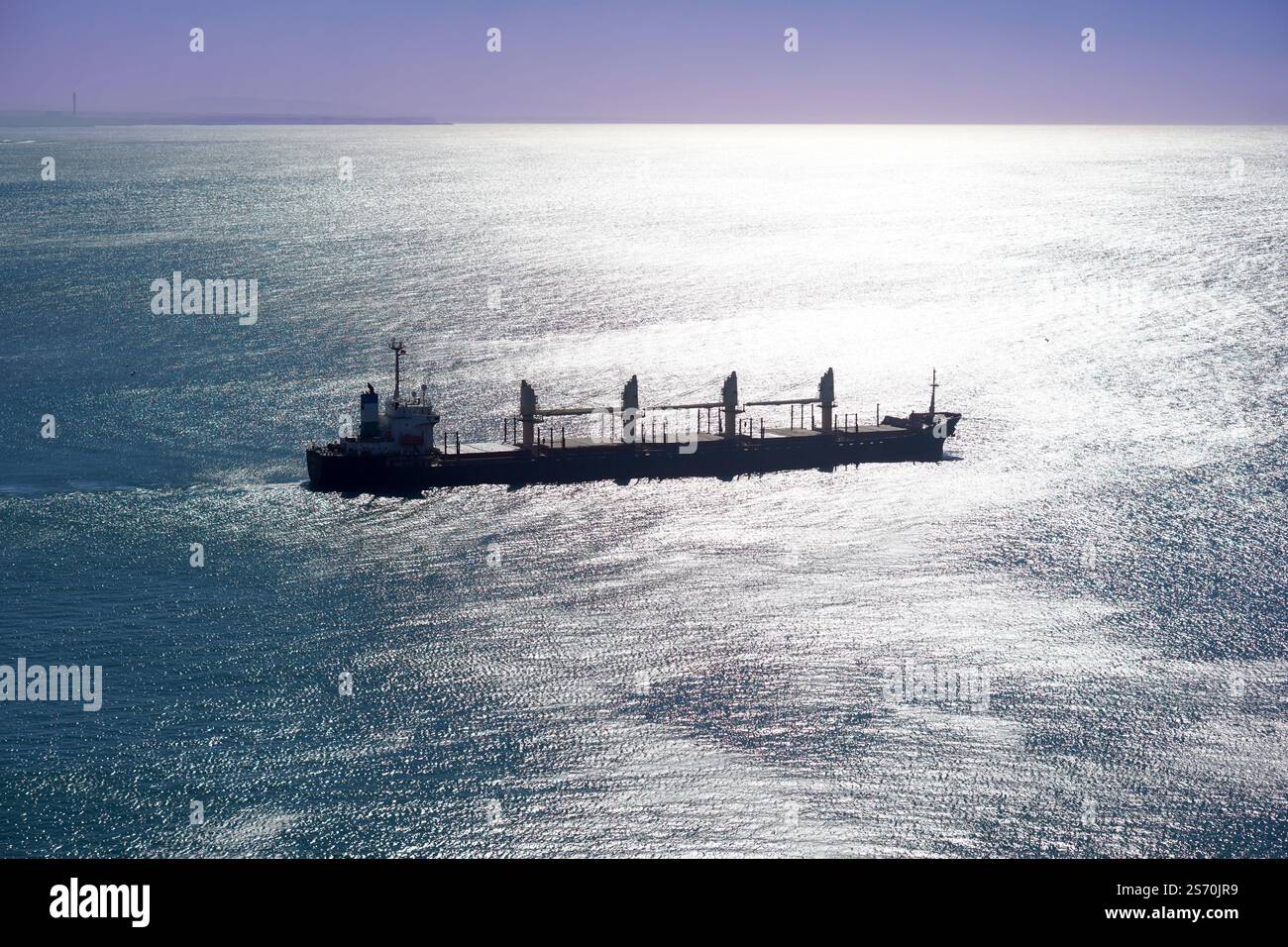 una grande nave da carico naviga su un mare scintillante sotto un cielo limpido. La luce del sole che si riflette sull'acqua mette in risalto la silhouette della nave Foto Stock