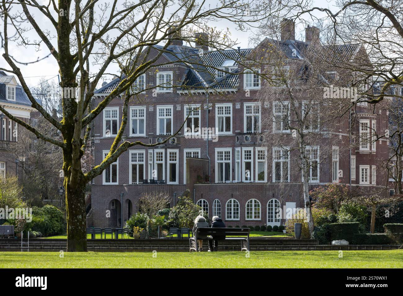 Edificio vicino al Vondelpark con 2 persone anziane su una panchina nell'erba verde lungo l'acqua in inverno, Amsterdam Paesi Bassi. 27 marzo 2023. Foto Stock