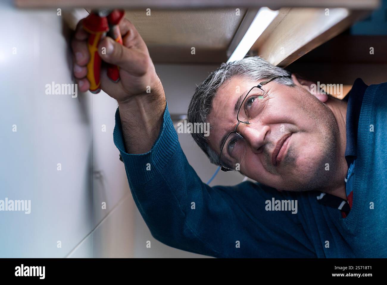 Man si è concentrato sulla riparazione di impianti elettrici sotto un armadio utilizzando delle pinze, eseguendo interventi di manutenzione precisi. Foto Stock