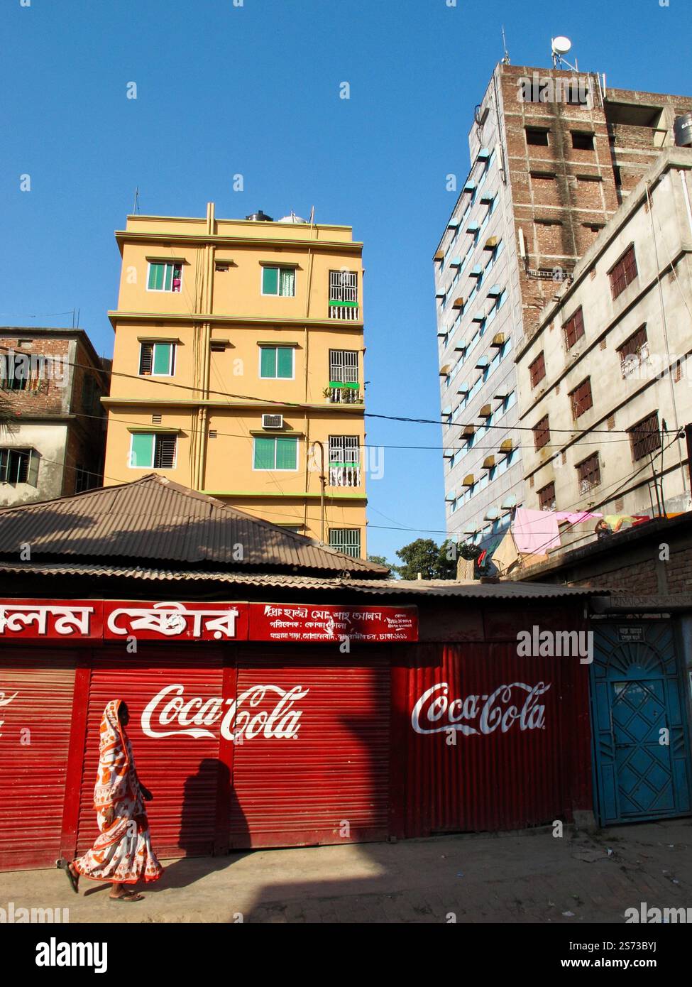 Una donna in un sari passa davanti a un cartello Coca-Cola nella Vecchia Dacca, Bangladesh, incorniciato da colorate architetture urbane. Foto Stock
