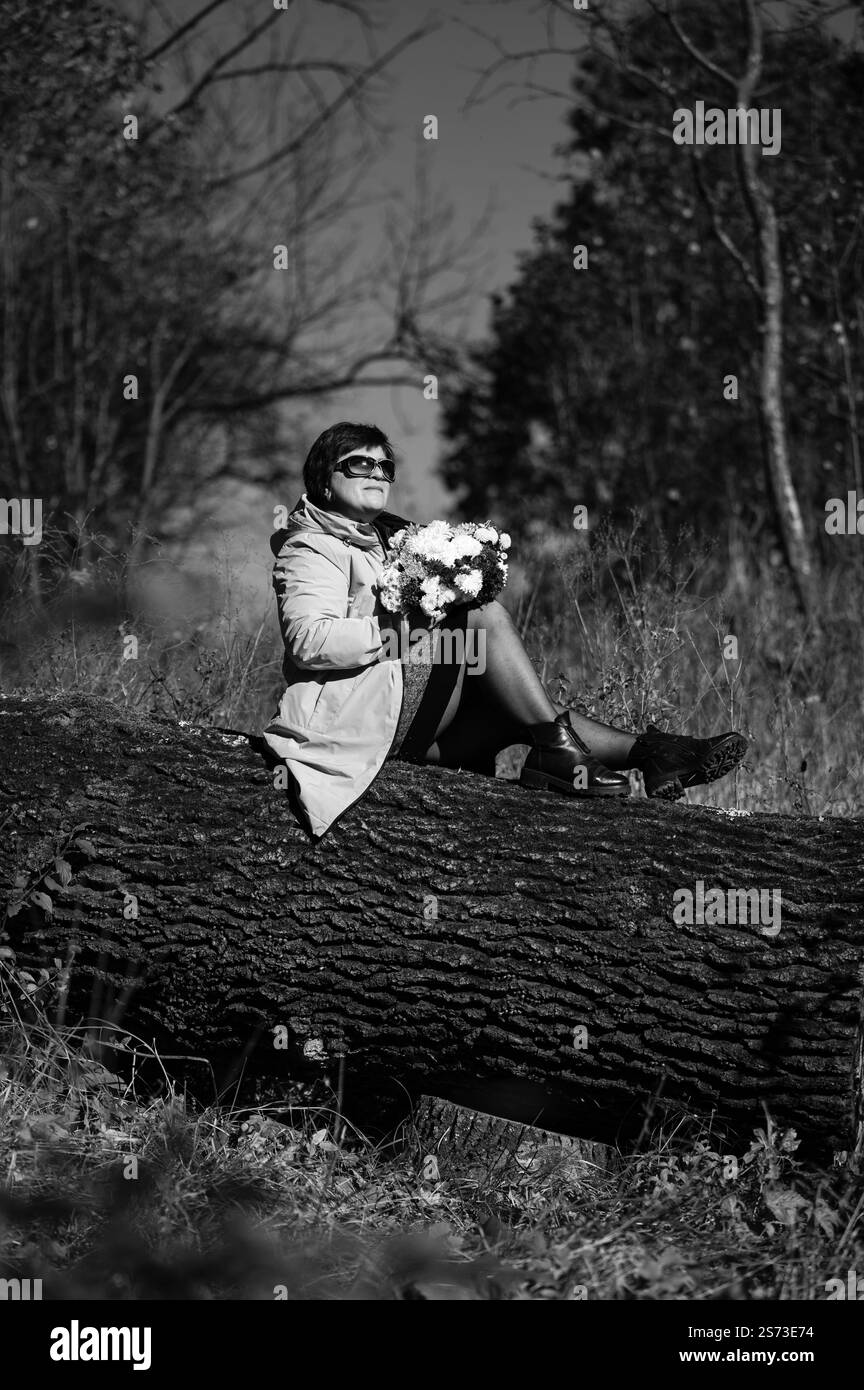 Una donna matura, che indossa un cappotto e occhiali da sole, siede su un tronco caduto in un parco, tenendo in mano un delizioso bouquet di fiori. La scena è tranquilla e serena. Foto Stock