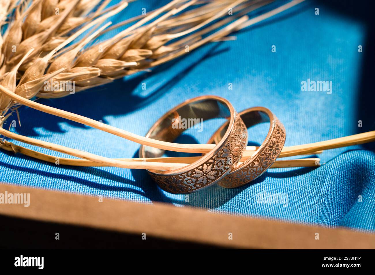 Due fasce nuziali decorate in oro poggiano su uno sfondo in tessuto blu, accompagnate da gambi di grano. La scena evoca temi di impegno e prosperità. Foto Stock