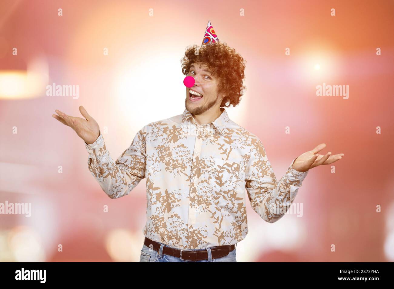 Ritratto di un giovane ragazzo felice con i capelli ricci che indossano un naso da clown. Luci rosse lampeggianti sullo sfondo. Foto Stock