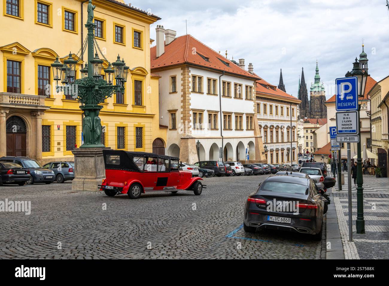 Una strada acciottolata di Praga con un'auto rossa e nera d'epoca, edifici storici gialli, auto moderne parcheggiate e la cattedrale di San Vito sul retro Foto Stock