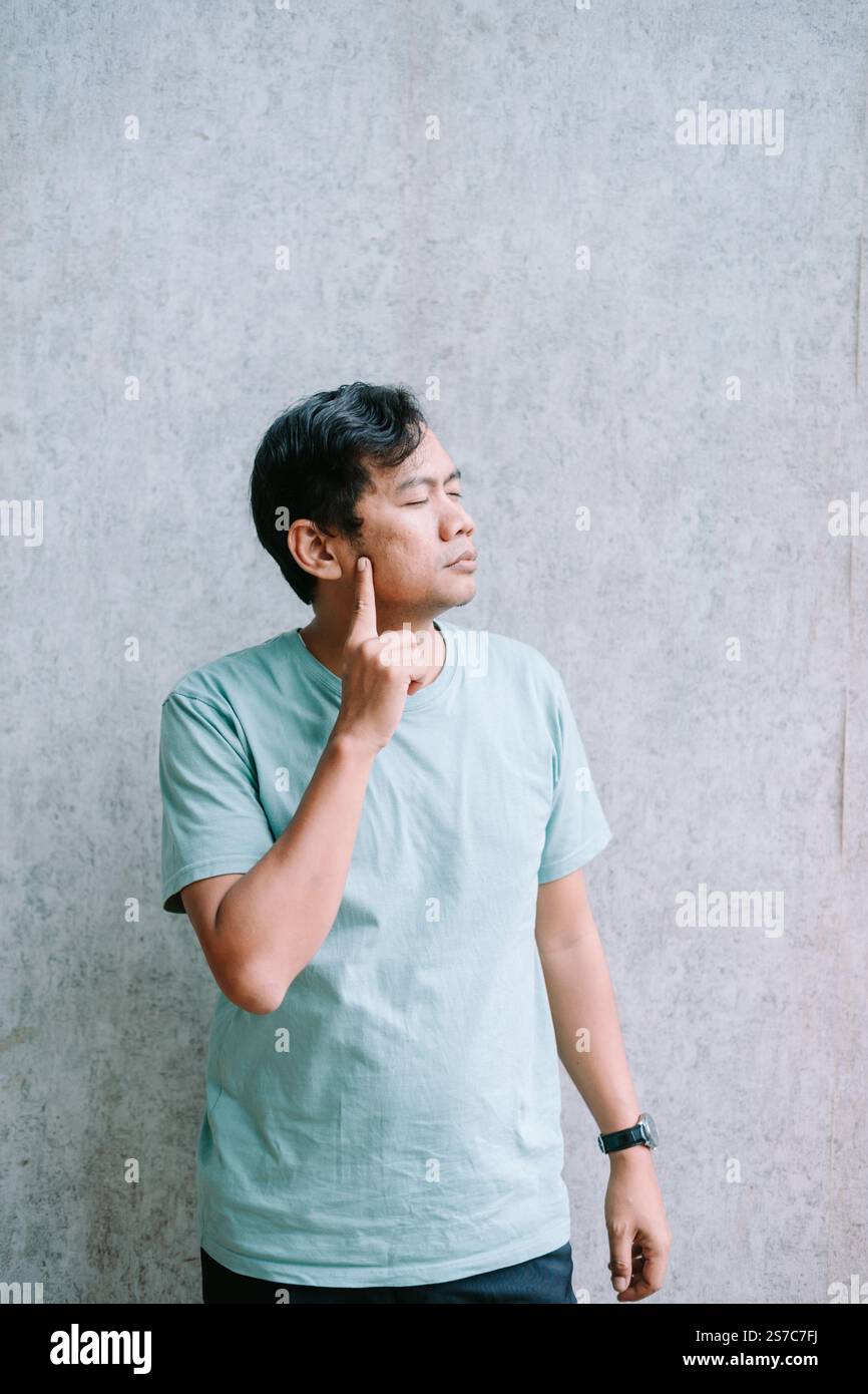 Un uomo con una camicia azzurra si staglia contro un muro testurizzato, puntando la sua guancia con un'espressione premurosa, trasmettendo un momento di profondo disprezzo Foto Stock